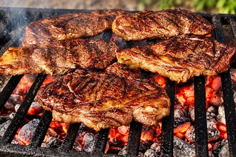 Beef steaks cooking over glowing charcoal on an iron grill.